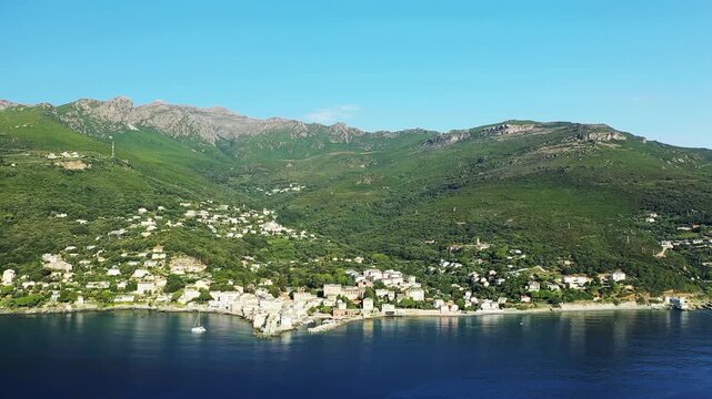 Scenic aerial shot of Erbalunga village nestled between lush green hills and the calm Mediterranean Sea on a clear day. Picturesque coastal landscape with traditional buildings and serene waters.
