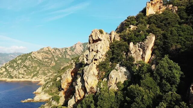Dramatic rocky cliffs and lush green vegetation at Calanques de Piana, Corsica, under a clear blue sky. The rugged coastal landscape overlooks the Mediterranean Sea.