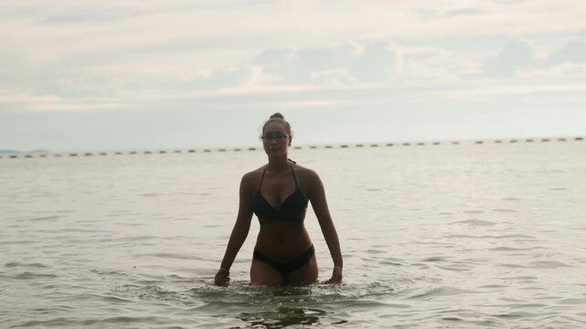 Black woman wading in ocean, confident model and fitness athlete, playful swimmer creating gentle splashes in shallow water, sunlit horizon and soft sky, empowered posture and graceful movements,