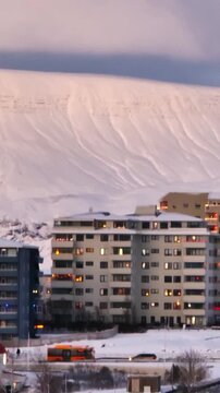 Reykjav&iacute;k apartment buildings in winter with glowing windows and snowy Mount Esja rising behind the city in soft evening light.