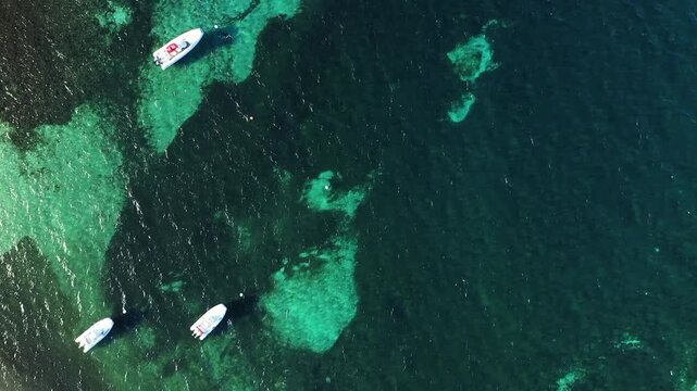Drone shot of boats floating on clear turquoise water near Plage du Petit Sperone, Corsica. The vibrant sea and scattered vessels create a tranquil coastal scene.
