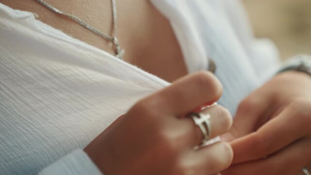 Closeup hands buttoning white shirt sunlit beachside scene, woman in black bikini, delicate silver rings and necklace, watch on wrist, textured lightweight fabric, intimate wardrobe routine, warm