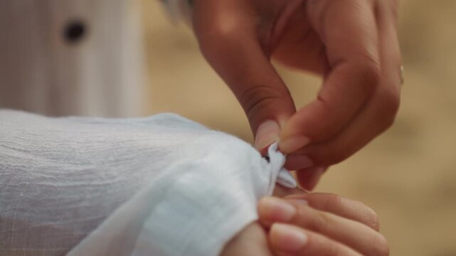 Closeup hands adjusting white linen sleeve on beach at golden hour, fingers rolling cuff to reveal bracelet and watch, gentle preparation ritual scenes suggest bride getting ready, sailor checking