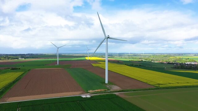 Aerial view of wind turbines generating renewable energy in the green and yellow patchwork fields of Normandy, France under a partly cloudy sky.
