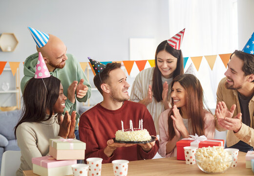 Joyful group of diverse young friends celebrating birthday at home with gifts and decorations. Smiling birthday person wearing party hat holding cake with candles while friends clapping and cheering.