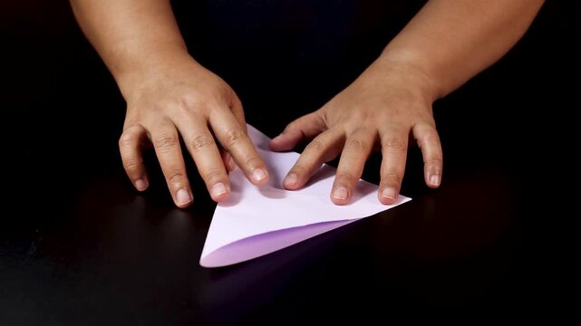 Hands folding purple paper into origami triangle shape on black background.