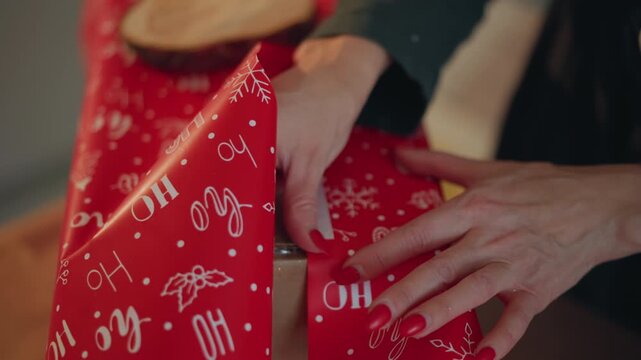 Closeup hands unwrapping red christmas present on table, warm living room lighting, careful fingers smoothing patterned paper and peeling seam, red nail polish, intimate holiday reveal by homeowner,