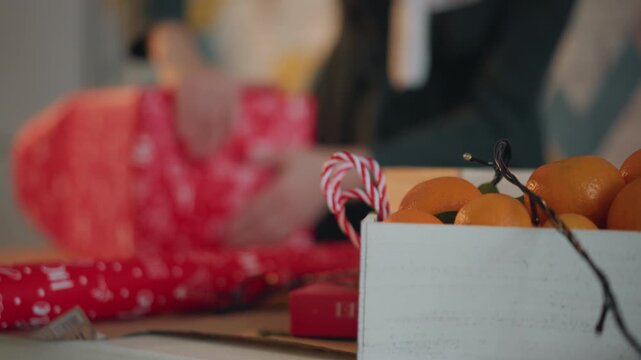 Hands wrapping red present near fruit crate, candy cane and oranges in wooden box, fairy lights and ribbon, busy festive table, hamper assembly, seasonal scent and joyful prep for christmas gifting