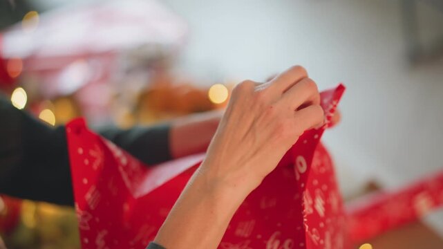 Closeup caucasian hands wrapping red present by tree with warm lights and bokeh, living room scene with festive ornaments, careful folding, tape and ribbon, intimate family atmosphere and joyful