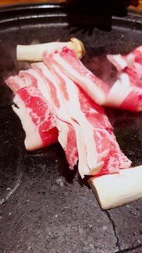 Vertical video. Thin Slices of Raw Beef Laid on Hot Grill Beside Mushrooms and Onion While Tongs Arrange Meat Under Bright Warm Light