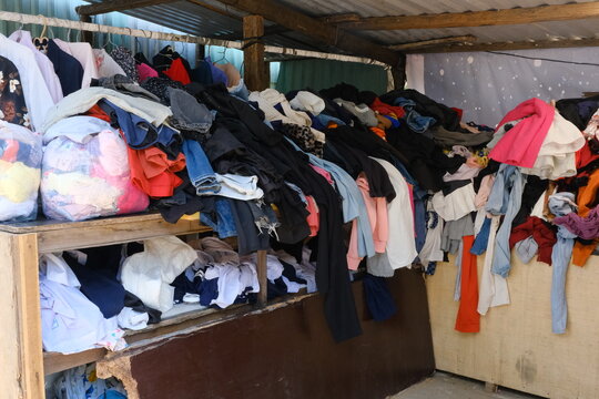 A cluttered display of assorted clothing piled on wooden shelves in a market setting. Various colors and styles are visible, creating a vibrant scene.