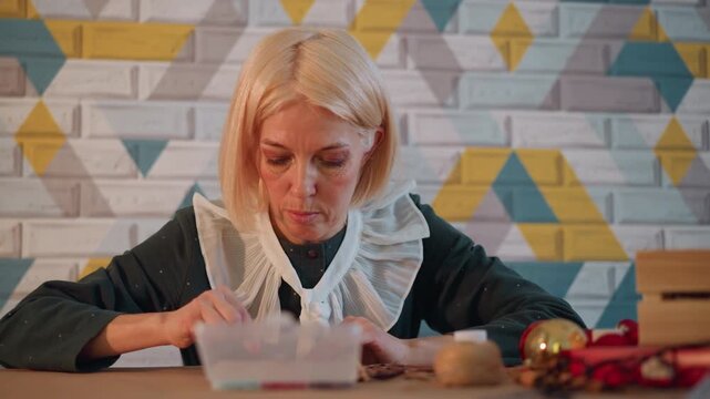 White woman wrapping small christmas present hands tying ribbon near ornaments twine wooden table patterned wall background concentrated expression careful craft warm fairy lights cozy holiday