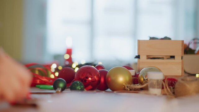 Christmas gift wrapping table with ornaments, hands placing ribbon on present, warm candlelight, wooden crate, red and gold baubles, scissors and twine, cozy bokeh background, festive atmosphere, home