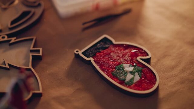 Closeup red mitten ornament on craft table, DIY crafter preparing Christmas present, glitter filling inside ornament, wooden tree cutouts scattered, scissors and twine nearby, warm bokeh lighting,