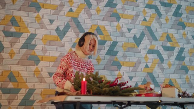 Cozy Christmas craft table, caucasian woman arranging wreath and wrapping present wearing headphones and festive sweater candles, ribbon, scissors and small gift box on table geometric patterned brick