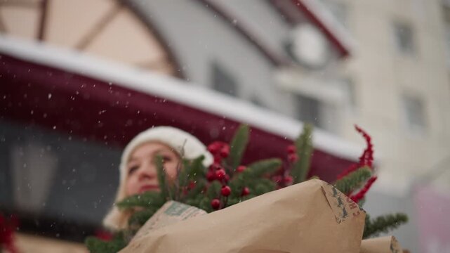 White woman holding christmas present bouquet in snow, urban market backdrop, green puffer coat and knit beanie, red berries and evergreen sprigs wrapped in kraft paper, smiling shopper pauses