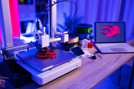 A 3D printer is actively creating a red object on its build plate, with filament spools and a laptop displaying a 3D model in the background