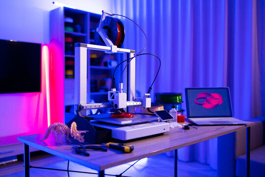 A 3D printer is actively creating a red object on its platform, with filament spools and a laptop displaying a 3D model nearby