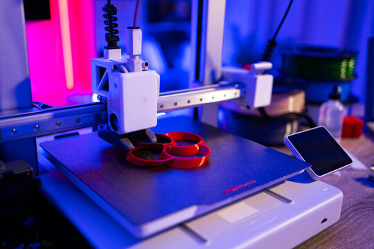 A modern 3D printer is actively creating a red object layer by layer on its build plate with vibrant blue and pink lighting