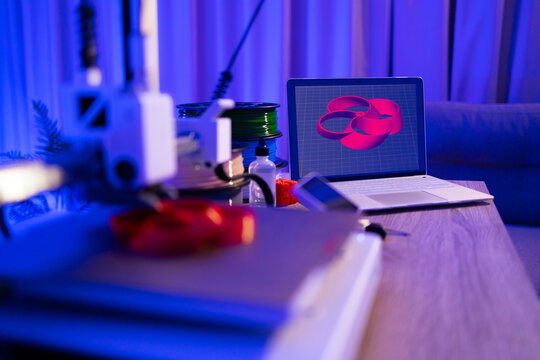 A 3D printer is set up next to a laptop displaying a 3D model, with spools of filament nearby, creating a modern maker space
