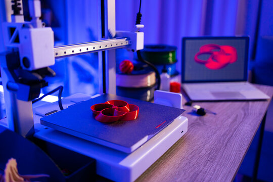 A 3D printer is actively creating a red, intricate object on its build plate, with filament spools and a laptop displaying a 3D model in the background