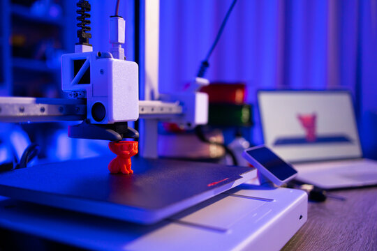 A 3D printer is actively creating a small orange object on its build plate, with a laptop displaying the design in the background