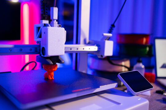 A 3D printer is actively creating a small orange figurine on its build plate, illuminated by vibrant blue and pink neon lights
