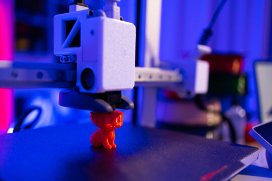 A close-up shot of a 3D printer in action, meticulously crafting a small, bright red cat figurine layer by layer under vibrant blue and pink lighting
