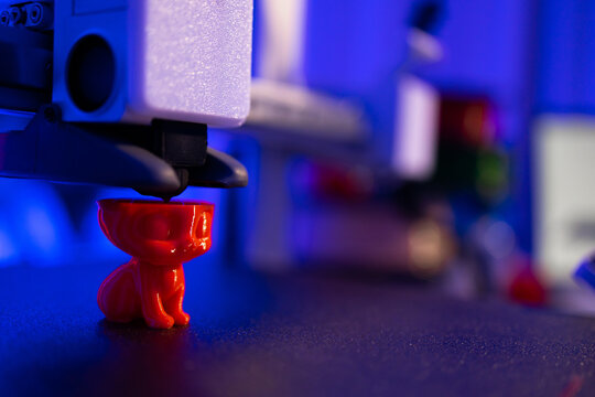 A 3D printer nozzle deposits molten plastic layer by layer to create a small, bright red cat figurine in a dimly lit workshop