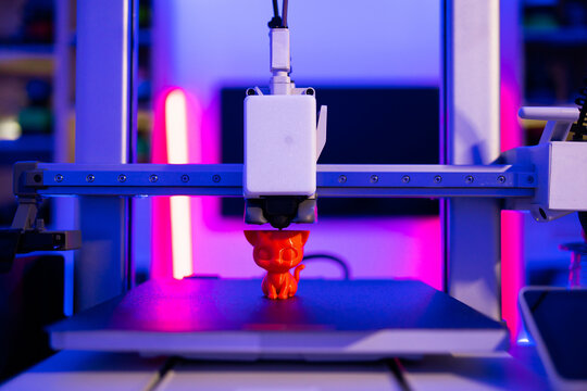 A 3D printer is actively creating a small, orange cat figurine with intricate details under vibrant blue and pink neon lighting
