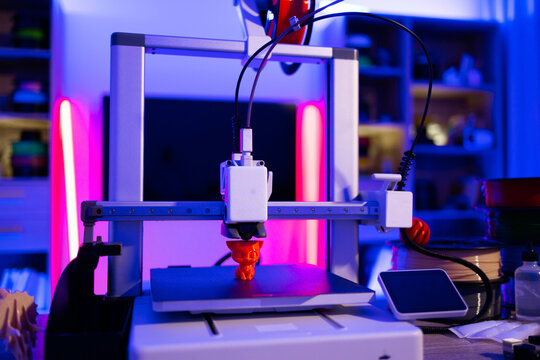 A modern 3D printer is actively creating an orange figurine on its build plate, illuminated by vibrant neon lights