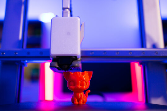 A 3D printer is in the process of creating a small, bright orange cat figurine with intricate details