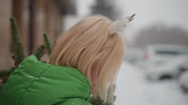 Blonde white woman carrying small tree, turning head while walking on snowy sidewalk in green coat with reindeer ear headband, wrapped gift box in basket, soft snowfall, nostalgic holiday return scene