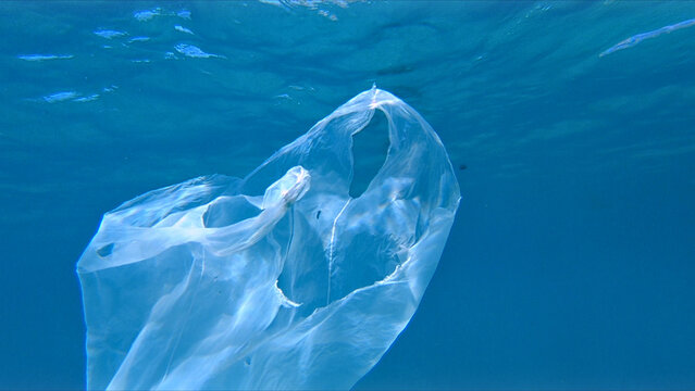 A large transparent plastic bag floats in clear blue water, highlighting environmental damage. The plastic, likely a bag, is suspended, emphasizing the pervasive issue of water pollution.