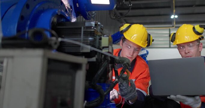 
Two industrial technicians in protective gear analyze data from a robotic arm using a laptop in a modern factory setting.
