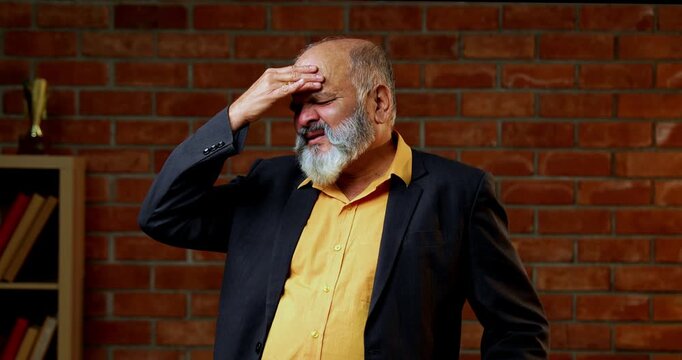 Indian senior businessman or corporate executive suffering from a painful headache and touching his temples while standing against a red brick wall in a cozy office with moody lighting.