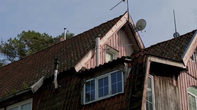 Old house with clay tile roof and dormer window. Weathered wooden exterior architecture of a vintage building. Residential dwelling with chimney and antenna against a clear sky.