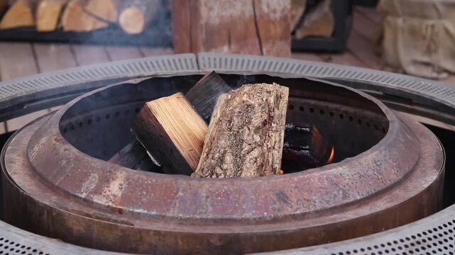 Wooden logs burn in a modern metal fire pit (pit ring) on ​​a wooden terrace. Setting up a fire pit on the street, in the garden or at the cottage. Fire in a metal fire pit outdoors. Coziness and rela