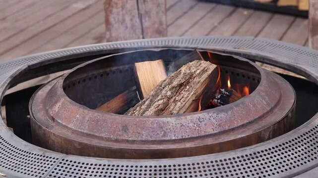 Wooden logs burn in a modern metal fire pit (pit ring) on ​​a wooden terrace. Setting up a fire pit on the street, in the garden or at the cottage. Fire in a metal fire pit outdoors. Coziness and rela