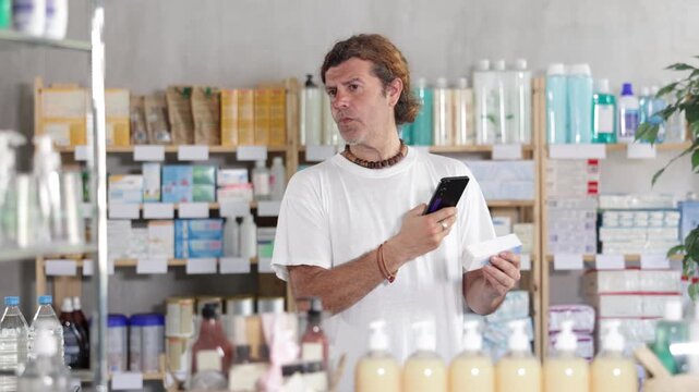 European man in summer clothes holds a mobile phone and scans the barcode on a package of ointment at a pharmacy. Buyer uses a non-cash method of payment for purchases. High quality 4k footage