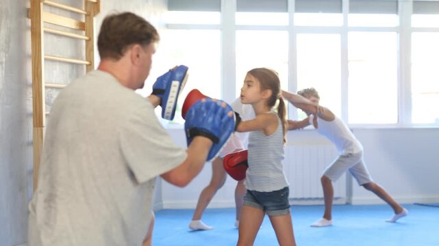 Girl and dad are boxing in gym, father helps daughter child to work out force of blow. Modern positive family chooses active hobby