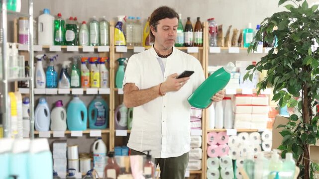 Thoughtful man buyer scans barcode using application on mobile phone, choose, liquid detergent. Customer follows link and reads information about product, data on recycling of packaging