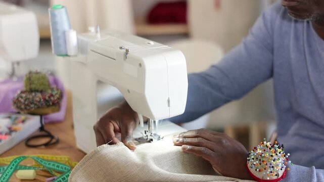 Close-up of hands of professional black male tailor working at sewing machine, stitching beige linen (soft lilac) fabric with precision at table in workshop