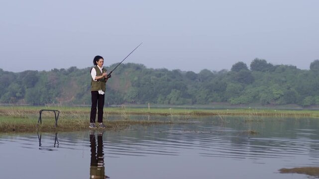 A South Asian girl wearing outdoor vest fishes quietly while standing alone in a lake, seen from behind