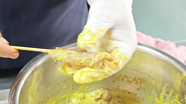 Preparing pork satay on skewers with yellow turmeric marinade in a metal bowl.
