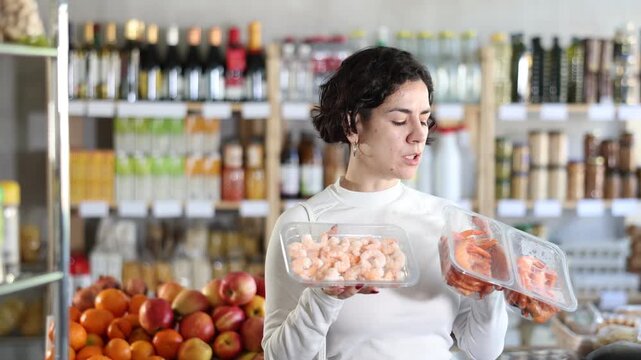 Woman choosing compare varieties cooled fresh shrimps, doubt and think before buying. Client choose and decide purchase prawns package, inspect good. Shop visitor pick up food in minimarket