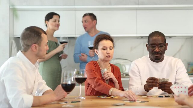 Joyful friends in their middle years sitting around table playing with cards and drinking wine in goblets