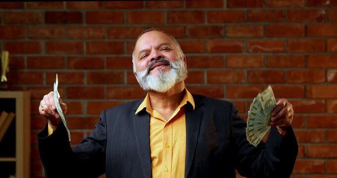 Indian senior businessman flaunting a fan of five hundred rupee notes to the camera with joy and success while standing against a red brick wall in a cozy office with professional moody lighting