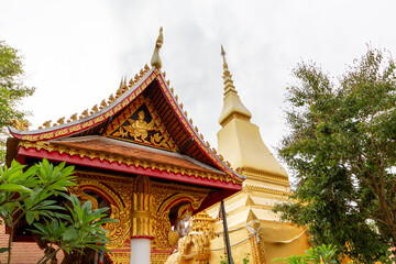 Fototapeta premium A beautifully decorated Thai temple pavilion with a red roof standing beside a magnificent golden stupa under a bright sky.