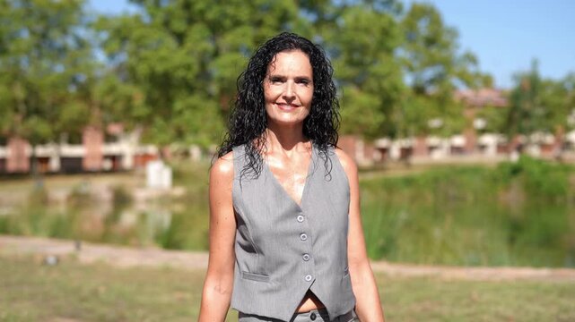 Woman smiles warmly outdoors. She gives a thumbs-up gesture. Natural daylight highlights her relaxed expression. She wears a simple gray vest top. Green trees and grass surround her